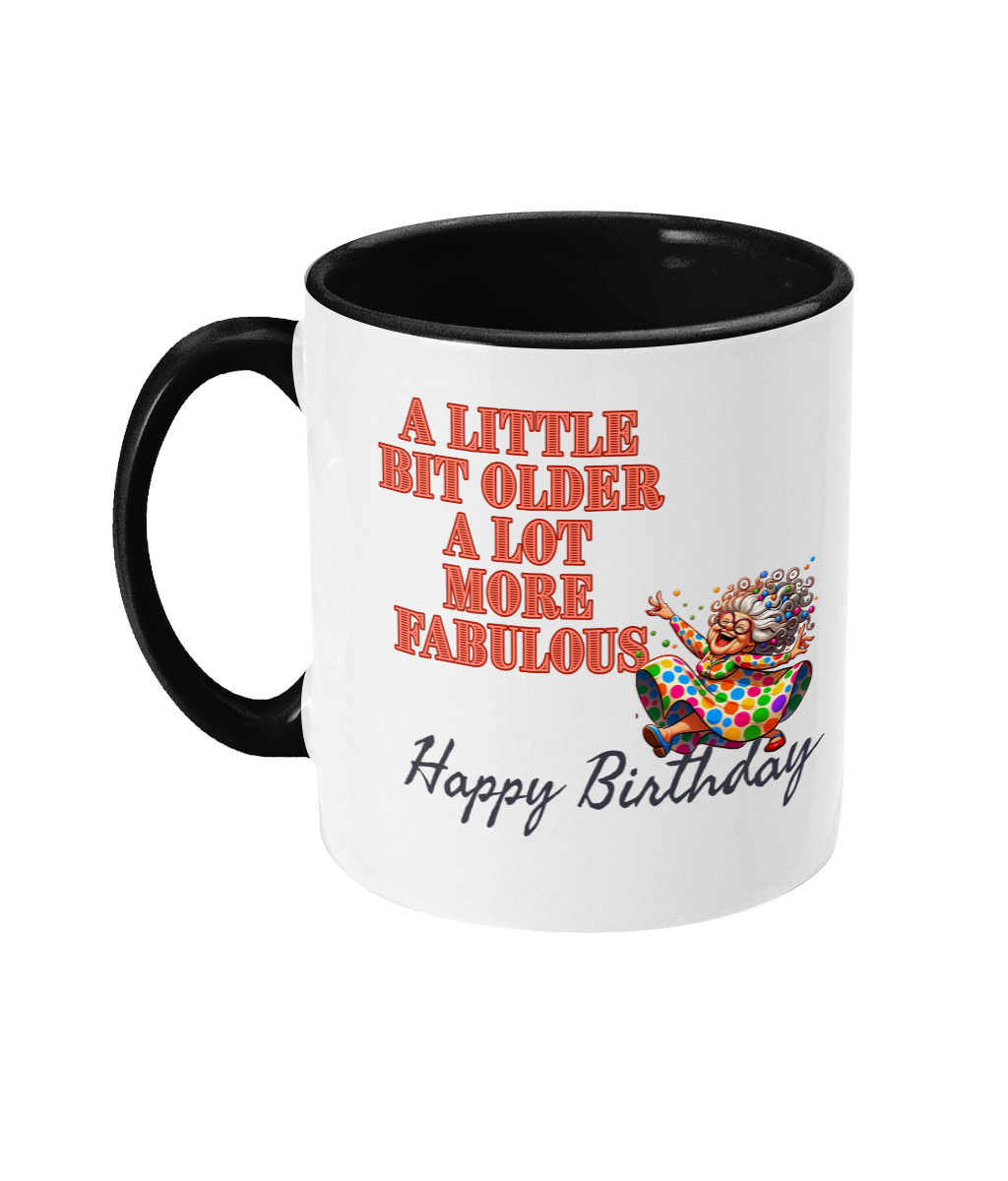 A white ceramic coffee mug with a black interior and handle, featuring colorful text and illustrations. The text reads, “A little bit older, a lot more fabulous” in bold red letters, followed by “Happy Birthday” in a cursive font. The design includes a cheerful illustration of a vibrant older woman in a colorful polka-dot dress, celebrating energetically.