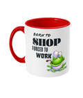 A white ceramic coffee mug with a red interior and handle, featuring the humorous text, “Born to shop, forced to work.” The design includes a colorful cartoon illustration of a happy green frog sitting on a lily pad, adding a playful and whimsical touch. The text is styled in bold fonts for emphasis.