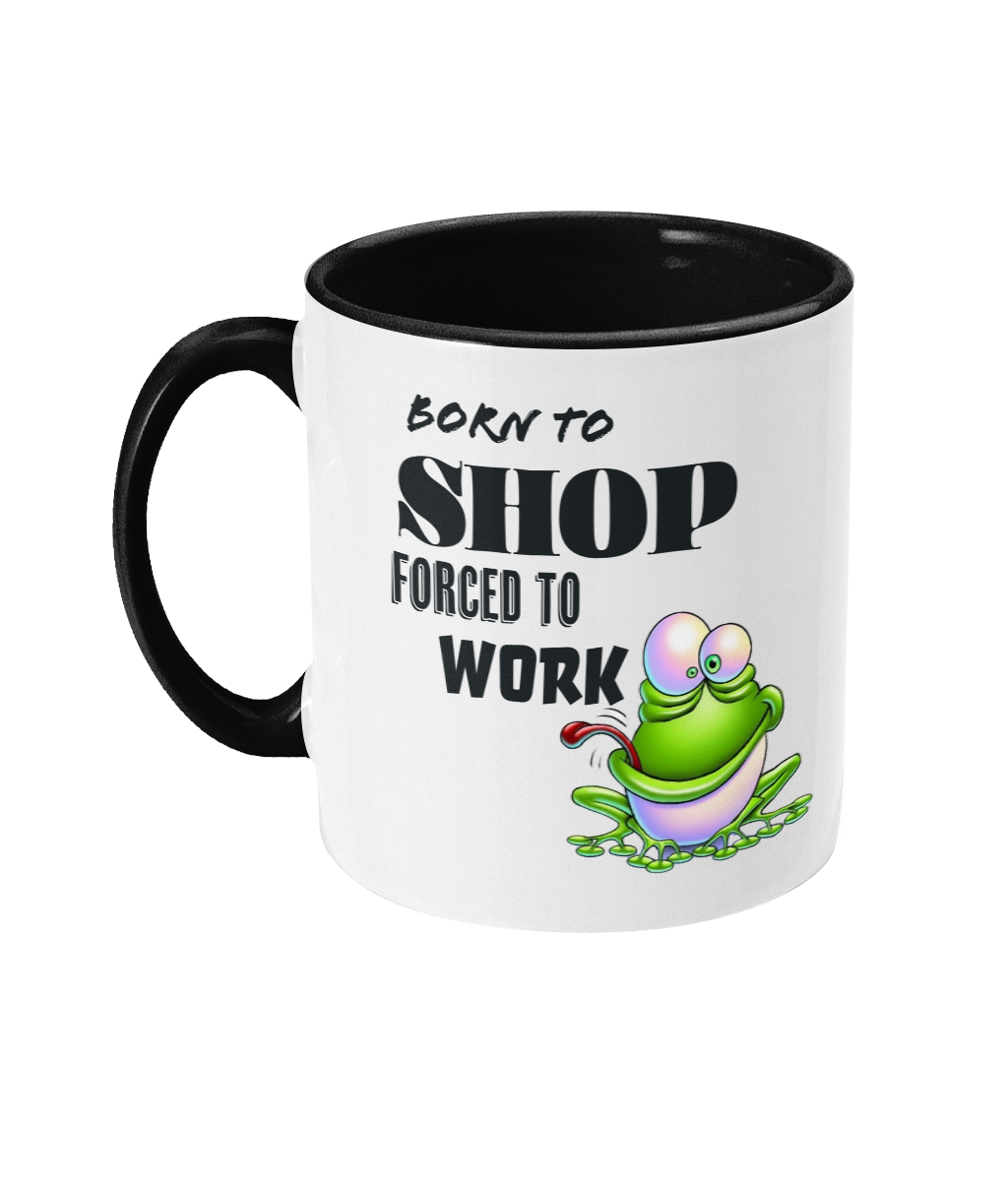 A white ceramic coffee mug with a black interior and handle, featuring the humorous text, “Born to shop, forced to work.” The design includes a colorful cartoon illustration of a happy green frog sitting on a lily pad, adding a playful and whimsical touch. The text is styled in bold fonts for emphasis.