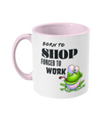 A white ceramic coffee mug with a pink interior and handle, featuring the humorous text, “Born to shop, forced to work.” The design includes a colorful cartoon illustration of a happy green frog sitting on a lily pad, adding a playful and whimsical touch. The text is styled in bold fonts for emphasis.