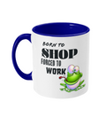 A white ceramic coffee mug with a blue  interior and handle, featuring the humorous text, “Born to shop, forced to work.” The design includes a colorful cartoon illustration of a happy green frog sitting on a lily pad, adding a playful and whimsical touch. The text is styled in bold fonts for emphasis.