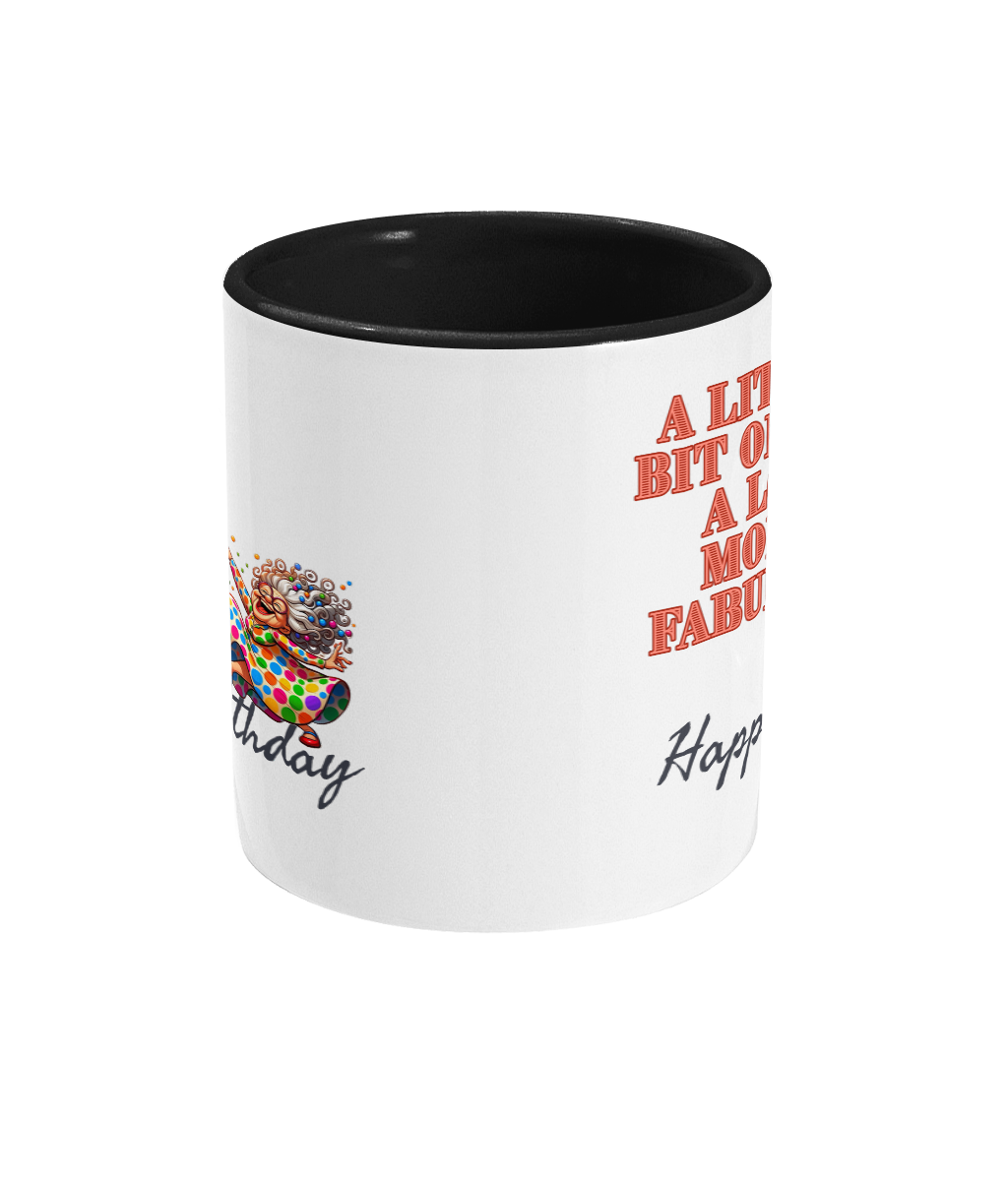 A white ceramic coffee mug with a black interior and handle, featuring colorful text and illustrations. The text reads, “A little bit older, a lot more fabulous” in bold red letters, followed by “Happy Birthday” in a cursive font. The design includes a cheerful illustration of a vibrant older woman in a colorful polka-dot dress, celebrating energetically.