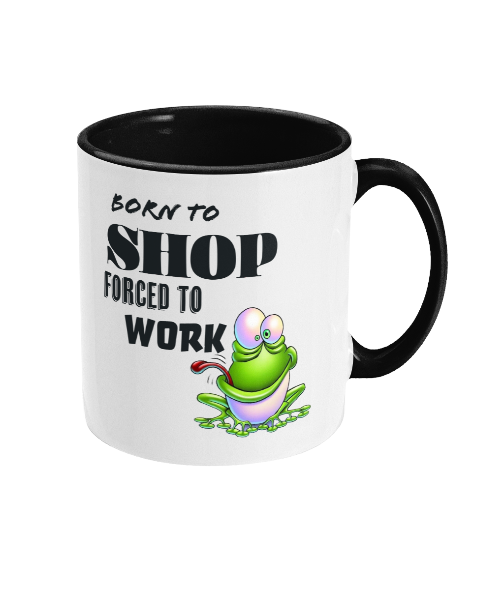 A white ceramic coffee mug with a black interior and handle, featuring the humorous text, “Born to shop, forced to work.” The design includes a colorful cartoon illustration of a happy green frog sitting on a lily pad, adding a playful and whimsical touch. The text is styled in bold fonts for emphasis.